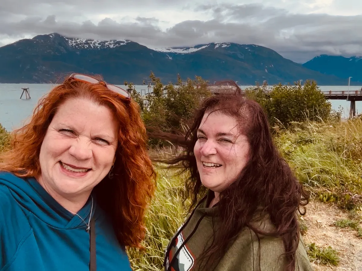 Heather Fulcher and Jody Fulcher Dennett, the sister-owners of Monroe Coffee Co., smiling together on the Alaskan coast with mountains in the background